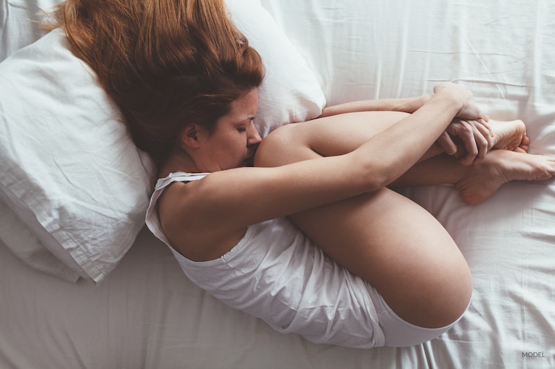 Woman curled up in bed, hugging her knees looking uncomfortable.